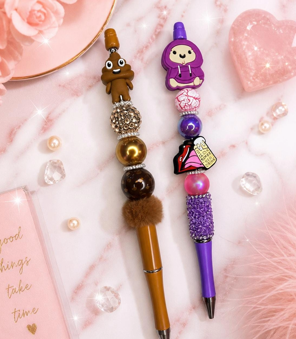 Two handmade beaded pens on a marble surface — one brown with poop emoji topper, one purple with hoodie character topper