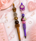 Two handmade beaded pens on a marble surface — one brown with poop emoji topper, one purple with hoodie character topper