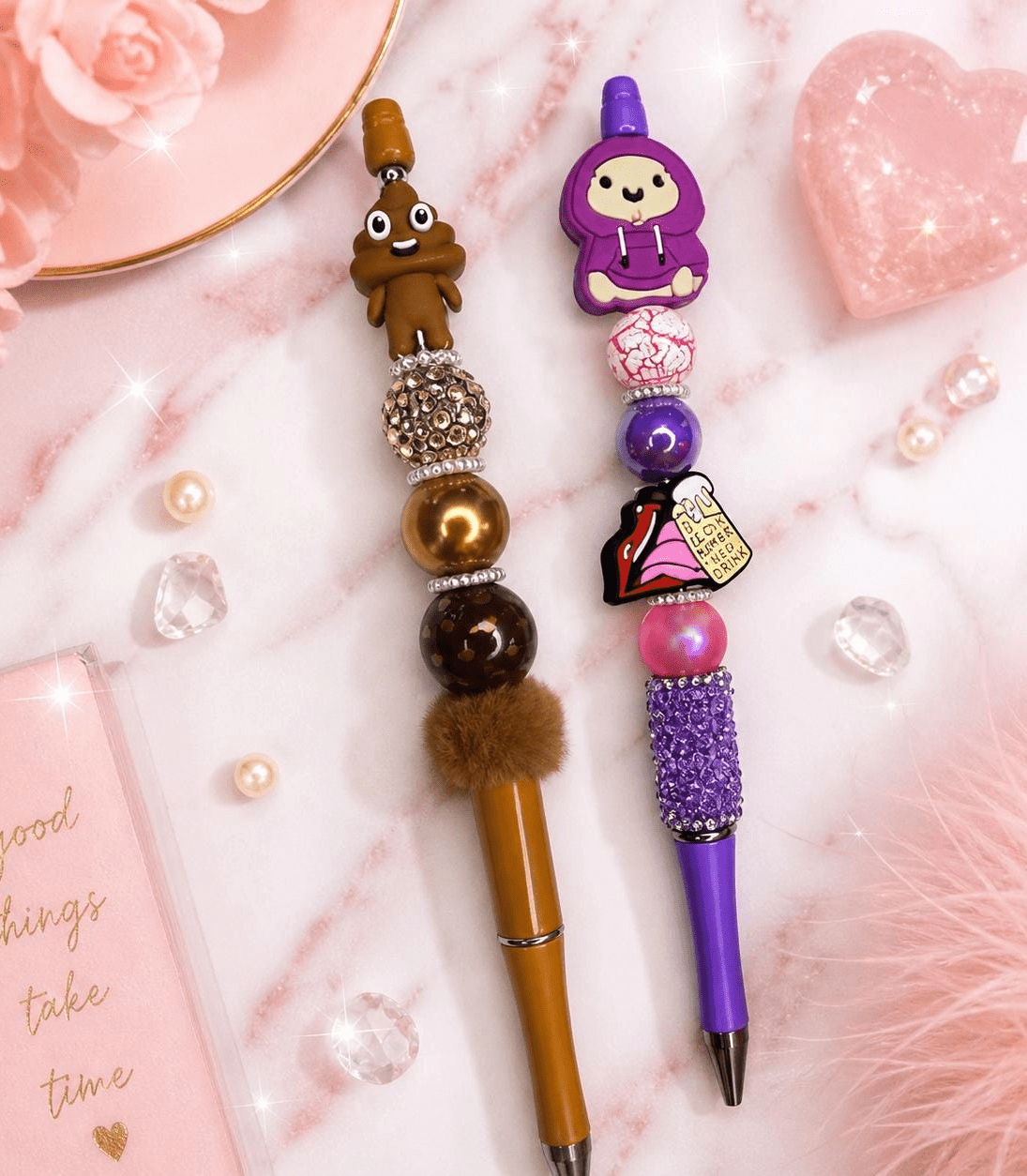 Two handmade beaded pens on a marble surface — one brown with poop emoji topper, one purple with hoodie character topper